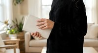 Woman in black knitted sweater holding white laptop computer in bright modern living room with plants and natural light for remote work and productivity.