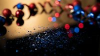 Festive blue glitter scattered on dark surface with red and blue Christmas ornaments and bokeh lights in background.