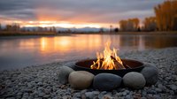 Cozy campfire burning in metal bowl surrounded by smooth stones on lakeside beach at sunset with autumn trees and mountain backdrop for outdoor recreation.