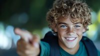 Young male with curly blonde hair and bright blue eyes smiling confidently at camera while pointing forward. Natural outdoor setting with soft bokeh background creates engaging portrait.