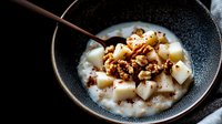 Creamy oatmeal porridge topped with diced pears, crunchy walnuts and spices in dark ceramic bowl. Moody lighting creates appetizing breakfast scene.