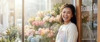 Young woman with wavy brunette hair and warm smile wearing cream cardigan and white apron standing in front of flower shop storefront with pink and blue hydrangeas, suitable for small business, retail, and entrepreneurship concepts.