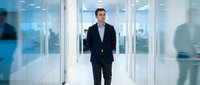 Confident businessman walking through modern office corridor with glass walls and blurred colleagues in background, corporate leadership concept.