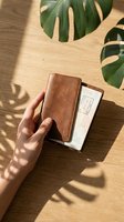 Person with medium skin tone holding brown leather wallet and white envelope on wooden surface with green monstera leaves and natural sunlight shadows.