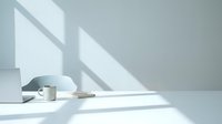 Minimalist white workspace with coffee cup and notebook bathed in natural window light creating geometric shadows on clean desk surface for productivity concepts.