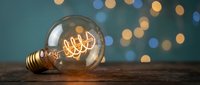 Vintage Edison bulb with glowing filament on wooden surface against teal background with warm bokeh lights for cozy interior design and lighting concepts.