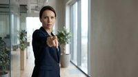 Professional businesswoman pointing finger at camera in modern office environment with confident expression and formal attire.
