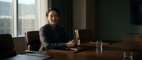 Professional Asian businessman in dark suit sitting at wooden conference table in modern office with city view through large windows during daytime meeting.