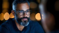 Middle-aged Indian businessman with salt and pepper beard wearing glasses and navy sweater, photographed in evening with soft bokeh lights in background.