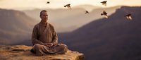 Young Asian monk in traditional robes meditating on mountain cliff at sunset with flying birds overhead creating peaceful spiritual atmosphere.