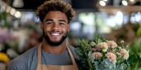 Young African American male florist with bright smile holding fresh floral arrangement in apron at flower shop, natural lighting and blurred background creates welcoming atmosphere.