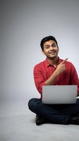 Young Indian man in red shirt pointing while using laptop computer sitting cross-legged on floor with excited expression for business and technology concepts.