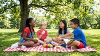 Diverse group of children enjoying outdoor picnic on checkered blanket under tree in sunny park during summer day.
