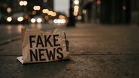 Cardboard sign with fake news text lying on urban street at night with blurred city lights creating bokeh effect in background for media concepts.