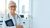Confident mature female doctor with gray hair and glasses smiling in modern medical office, holding tablet with brain scan for neurological consultation.