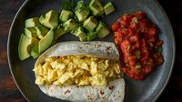 Breakfast burrito filled with scrambled eggs, topped with fresh diced avocado and homemade salsa on dark plate against rustic wooden background.