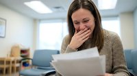 Young Caucasian woman in beige sweater covering mouth in shock while reading document in modern office waiting room with emotional reaction.