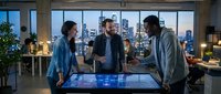 Three diverse business professionals collaborating around interactive digital table display in modern office with city skyline view at evening.
