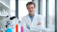 Confident middle-aged Caucasian male scientist in white lab coat standing with crossed arms in modern laboratory with microscope and equipment.