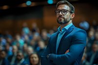 Confident businessman with glasses and beard in blue suit standing with arms crossed at professional conference with blurred audience background.