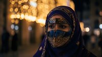 Middle Eastern woman wearing traditional ornate face veil and headscarf with intricate golden patterns against warm bokeh lights background at night.