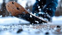 Close-up view of winter boots kicking up snow and ice crystals against blurred blue background, creating dynamic motion effect with scattered snowflakes in sunlight.