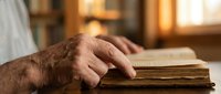 Elderly hands with age spots gently touching vintage book pages in warm golden library light creating peaceful reading atmosphere for seniors.
