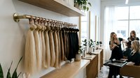 Hair extension samples displayed on wooden rack in modern salon with stylists consulting clients in background for beauty business.