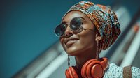Young African woman wearing colorful traditional headwrap and sunglasses with orange headphones around neck, smiling confidently in urban setting.