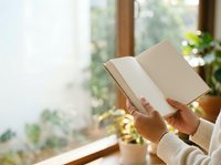Person with medium skin tone holding open blank notebook by sunny window with plants, ideal for education, learning, writing, journaling and lifestyle concepts.