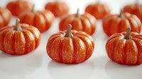 Small orange decorative pumpkins arranged in rows on white background, seasonal autumn gourds with natural stems and ribbed texture for harvest decor.