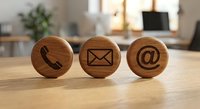 Three wooden circular tokens with engraved contact icons including phone, email and at symbol on office desk for business communication concepts.