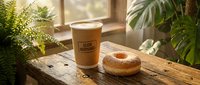 Takeaway coffee cup with glazed donut on wooden table surrounded by potted plants near window, morning breakfast or snack concept for cafes and food businesses.