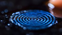 Concentric ripples in dark water create mesmerizing blue circles with bokeh light effects, capturing moment of impact and liquid surface tension dynamics.
