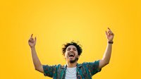 Young man with dark hair and beard, wearing casual patterned shirt, celebrating with arms raised and joyful expression against bright yellow background for success and happiness concepts.