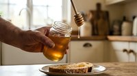 Closeup of male hand drizzling golden honey from wooden dipper onto whole grain toast on plate in bright kitchen, breakfast food preparation and healthy eating concept.