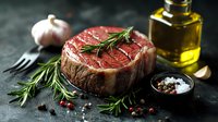 Raw beef tenderloin steak with fresh rosemary sprigs, garlic, olive oil, and seasoning on dark stone background, close up view of premium meat cut.