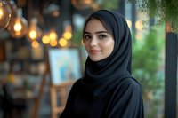 Young Middle Eastern woman wearing black hijab smiling warmly in modern cafe setting with soft bokeh lights and natural greenery creating welcoming atmosphere.