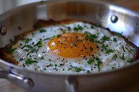 Fresh sunny side up egg cooking in stainless steel pan, garnished with chopped herbs and black pepper, showing vibrant orange yolk and crispy edges.