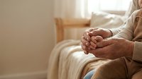 Senior woman sitting peacefully with clasped hands in warm sunlit room, expressing contemplation, prayer, and inner peace in cozy home setting.