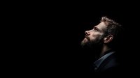 Profile view of bearded man in suit against black background, looking upward with contemplative expression, dramatic low-key lighting highlighting facial features.