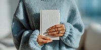 Woman in cozy blue sweater holding minimalist book with manicured hands and gold rings against soft blurred background.