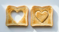 Two golden toasted bread slices with heart-shaped cutouts on white background, one with heart piece beside it, perfect for breakfast and love concepts.