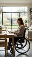 Woman in wheelchair working on laptop at wooden desk in bright home office with large windows overlooking garden, inclusive workspace design.