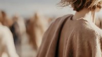 Back view of person with short hair wearing beige coat standing in crowd, soft focus background creates atmospheric depth for lifestyle and fashion content.