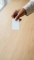 Middle-aged man with dark skin tone wearing beige linen shirt holding blank white business card over wooden table surface. Suitable for corporate branding and business identity mockups.