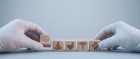 Hands in medical gloves arranging wooden blocks with healthcare symbols including stethoscope, heart, medical cross and pills on clean surface.