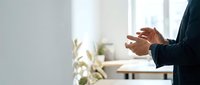 Professional businessman gesturing with hands during presentation or meeting in modern bright office space with natural lighting and plants.