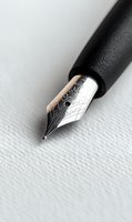 Close-up macro shot of fountain pen nib on textured white paper background, showing intricate metallic details and ornate engravings in silver and black tones.