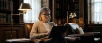 Mature woman with blonde hair and glasses working on laptop in traditional library setting with leather furniture and warm lighting.